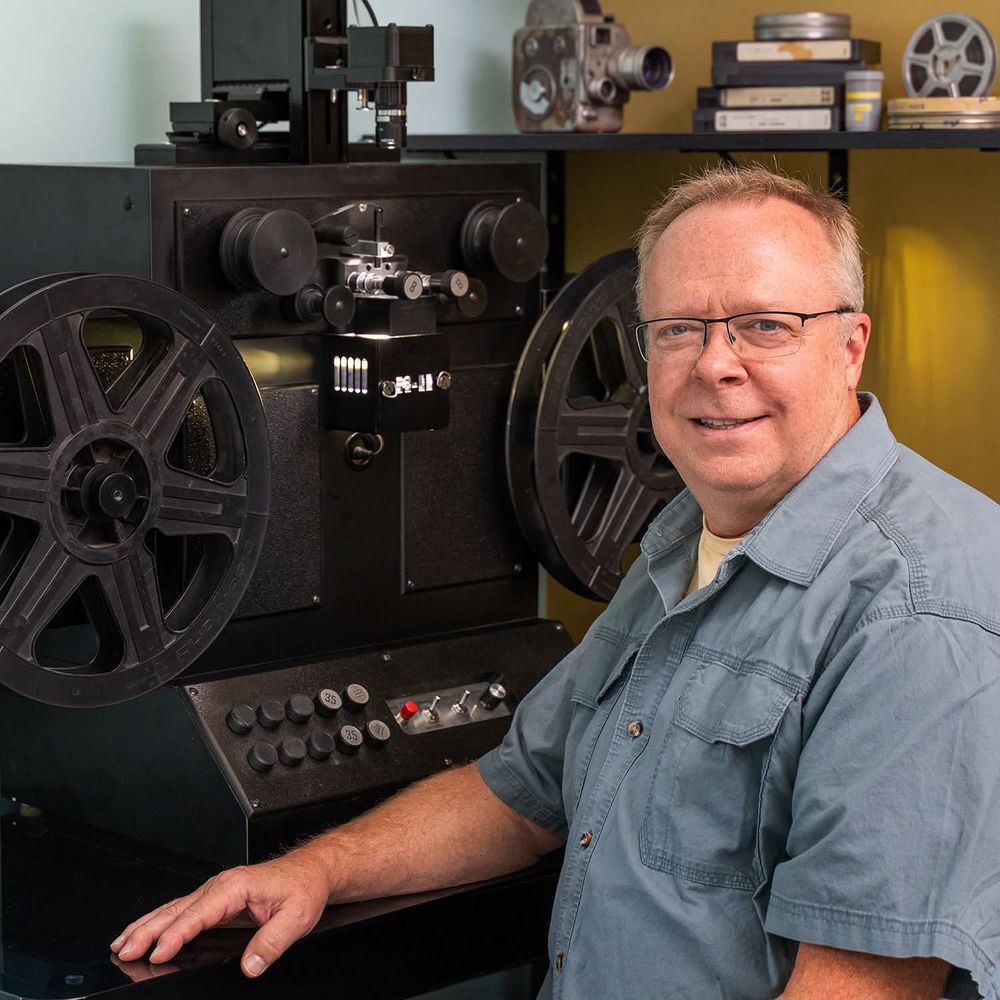 Dave Anderson seated beside his film scanner, with videotapes, film reels and a vintage movie camera on the shelf behind him